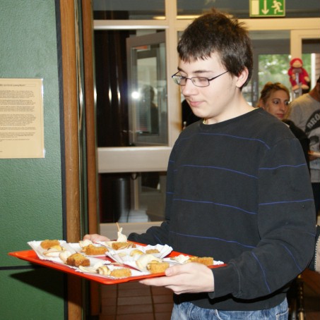 Ein Junge trägt ein Tablett auf dem kleine Pappteller sind. Auf jedem Teller ist 1 Glückskeks, 1 Stück Möhrenkuchen und 1 Rugelach.