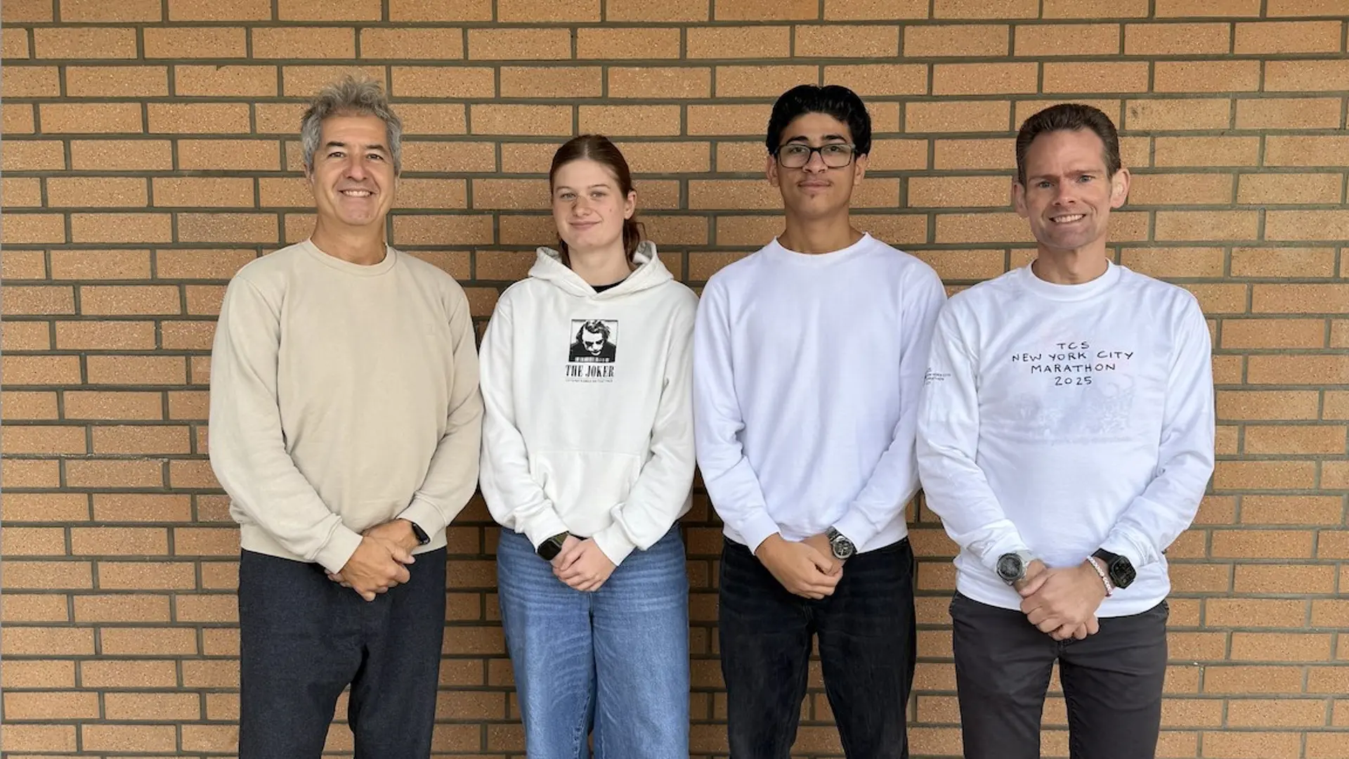 Vier Personen stehen nebeneinander vor einer braunen Klinkerwand: zwei Erwachsene außen und dazwischen ein Mädchen und ein Junge im Teenageralter.