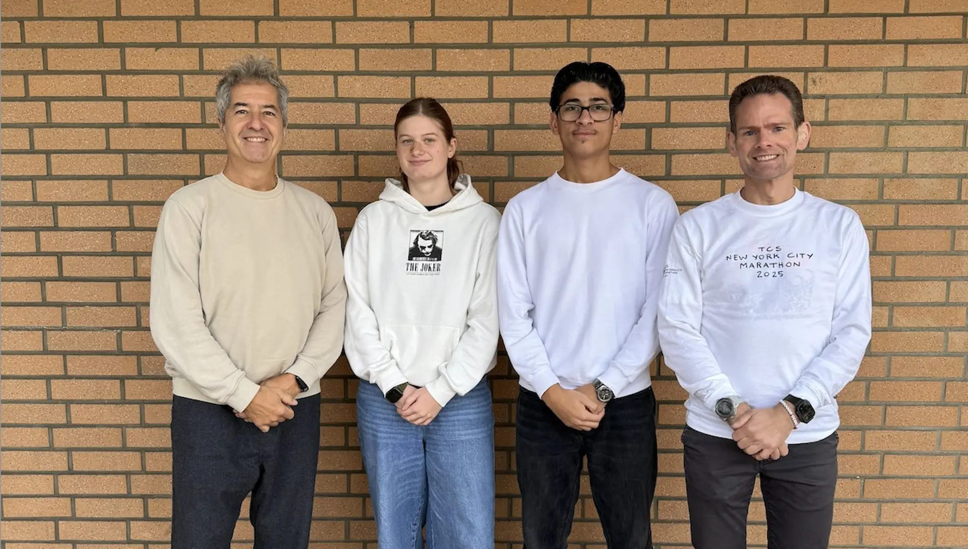 Vier Personen stehen nebeneinander vor einer braunen Klinkerwand: zwei Erwachsene außen und dazwischen ein Mädchen und ein Junge im Teenageralter.