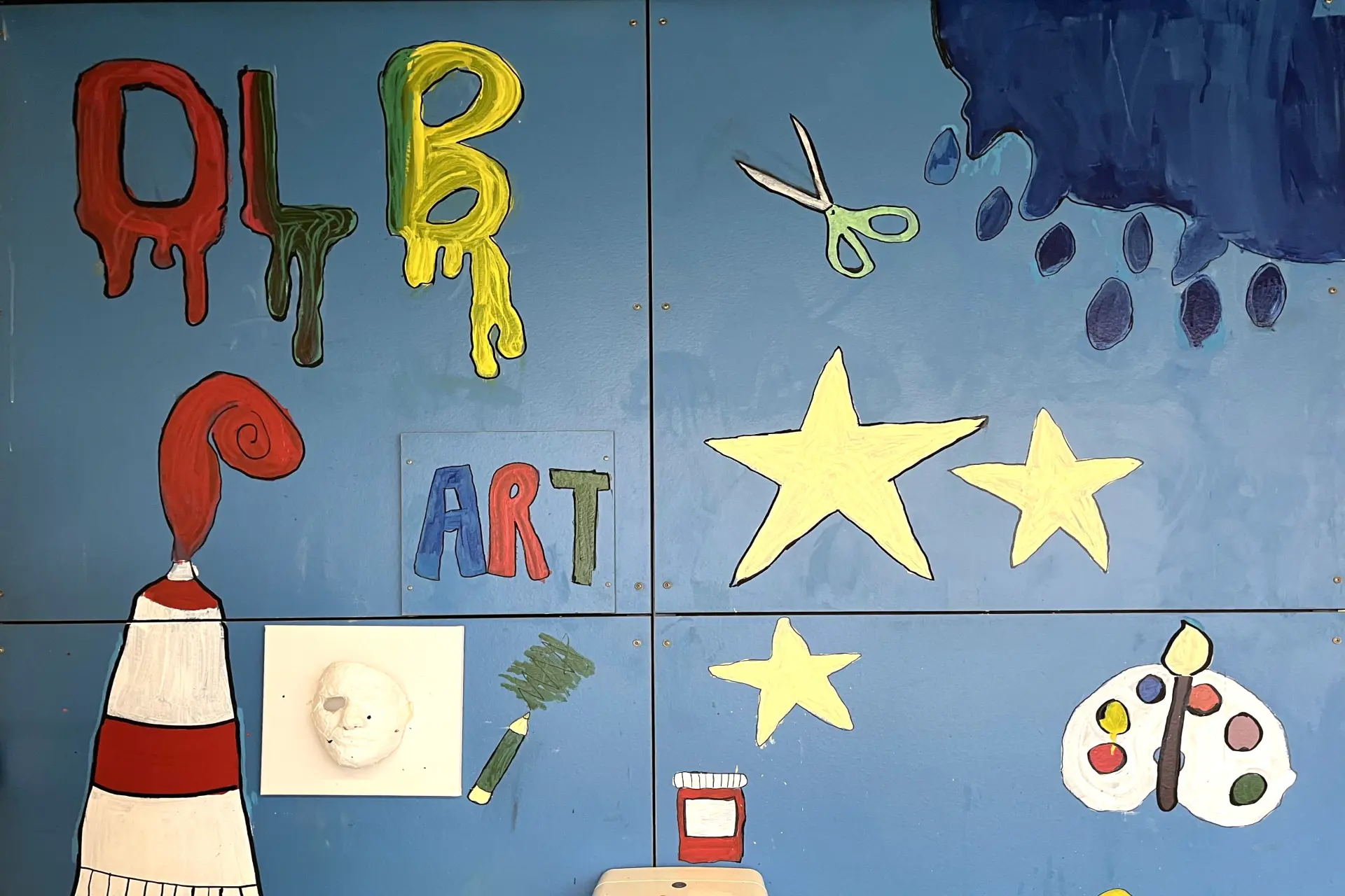 Eine hellblaue Wand mit verschiedenen handgemalten Kunstmotiven, darunter die tropfenden Buchstaben "DLB", das Wort "ART", eine Farbtube, Sterne und eine Malerpalette.