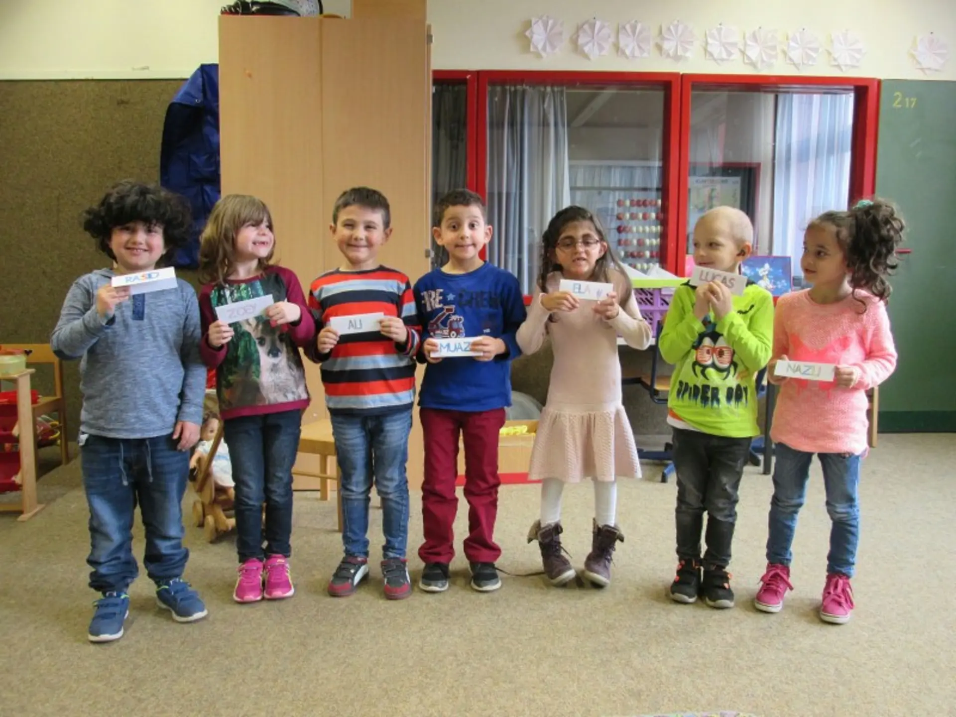 Sieben Kinder stehen in einem Raum. Jedes Kind hält einen kleinen Zettel mit dem eigenen Vornamen vor sich.