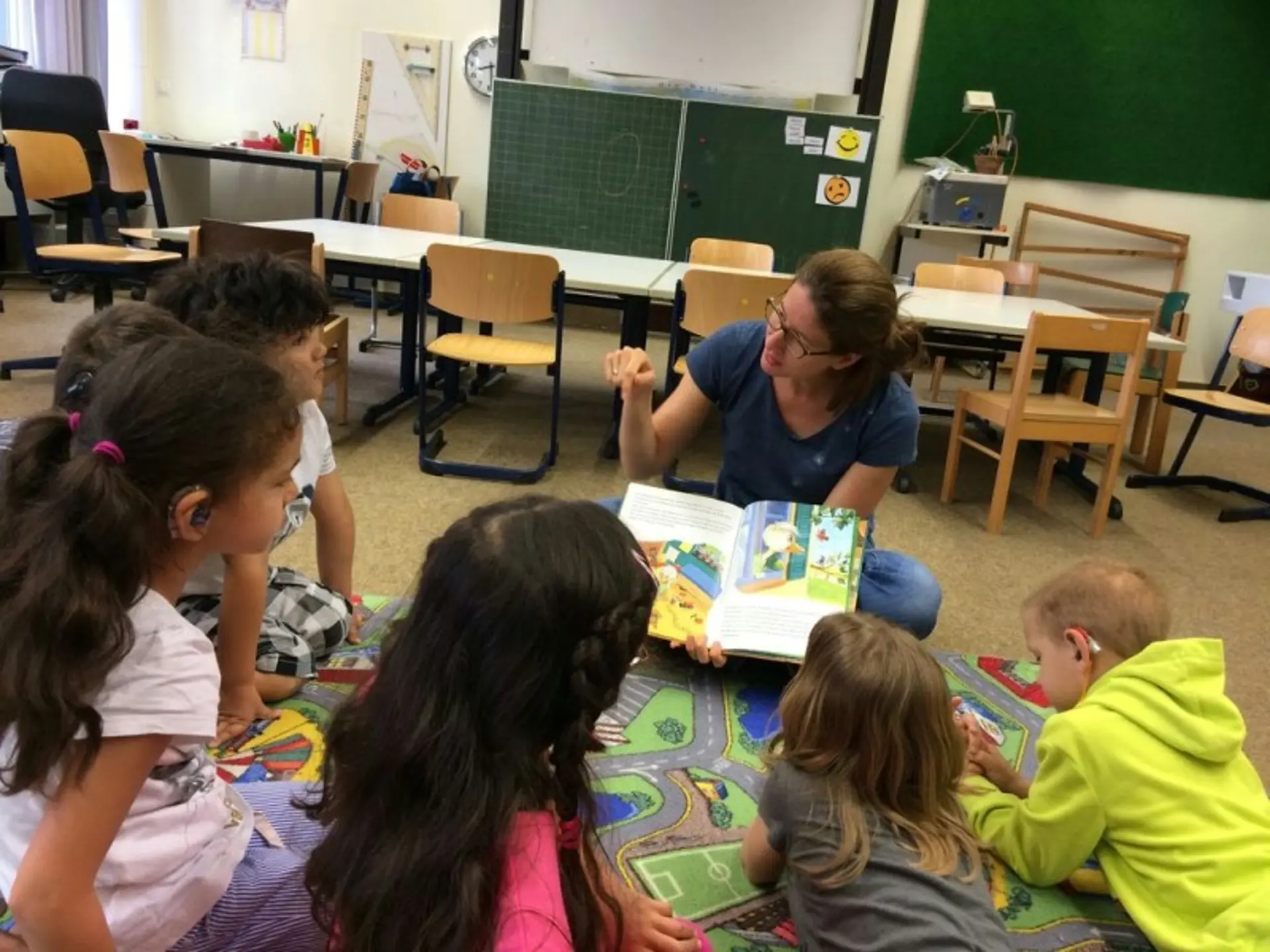 In einem Klassenzimmer sitzt einen Frau auf dem Fußboden. Vor ihr sitzen oder liegen sechs Kinder. Die Frau zeigt ein Buch und liest das Buch mit Handgebärden vor.